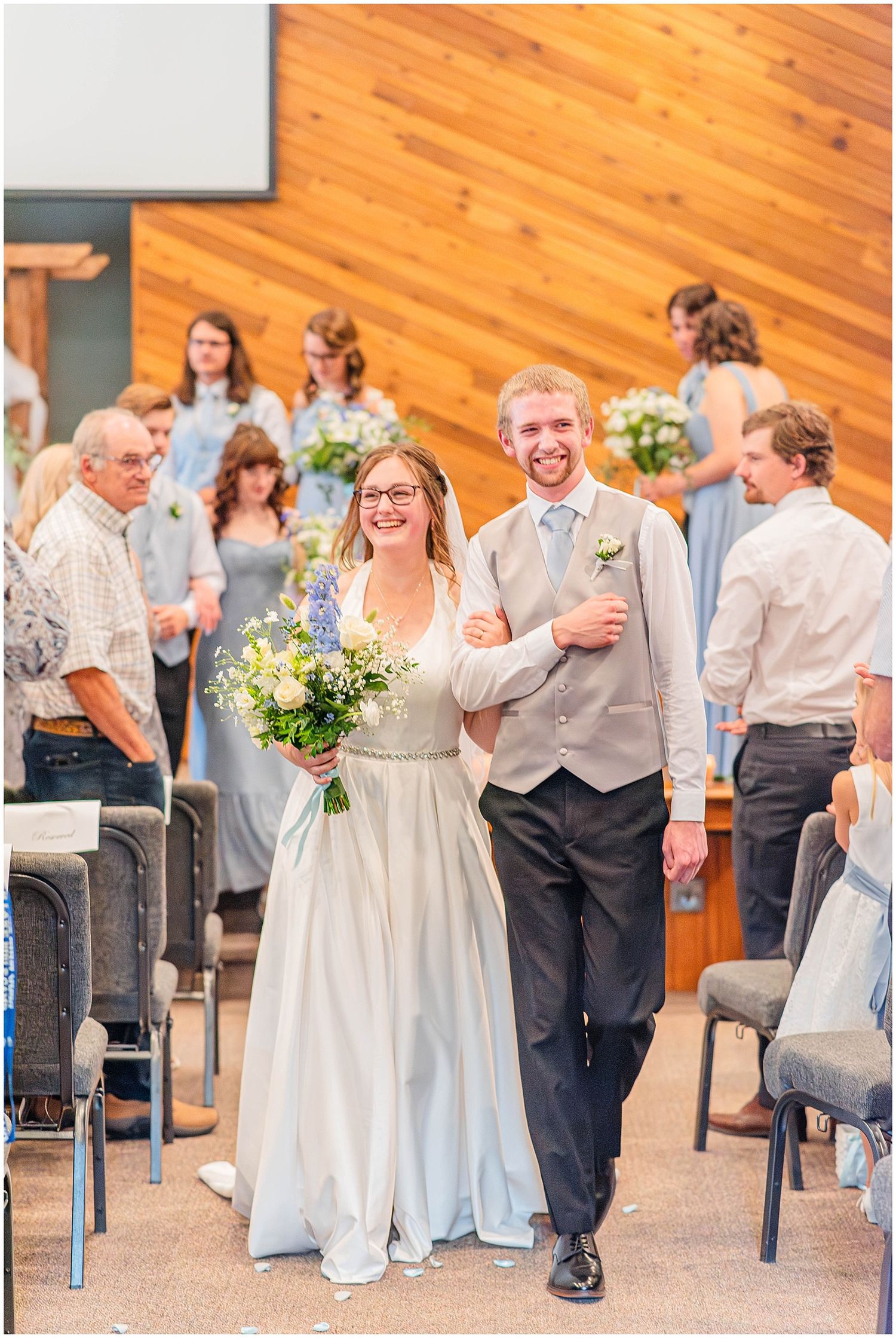 Smiling newlyweds walk down aisle together after wedding ceremony in church with bridal party behind them.