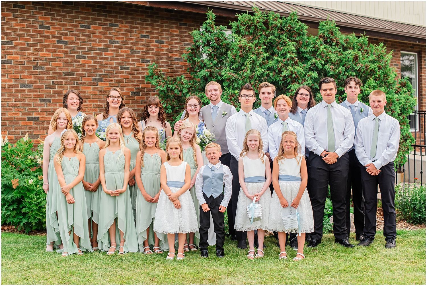 Large wedding party group photo with bridesmaids in sage green dresses and groomsmen in dark suits outdoors.