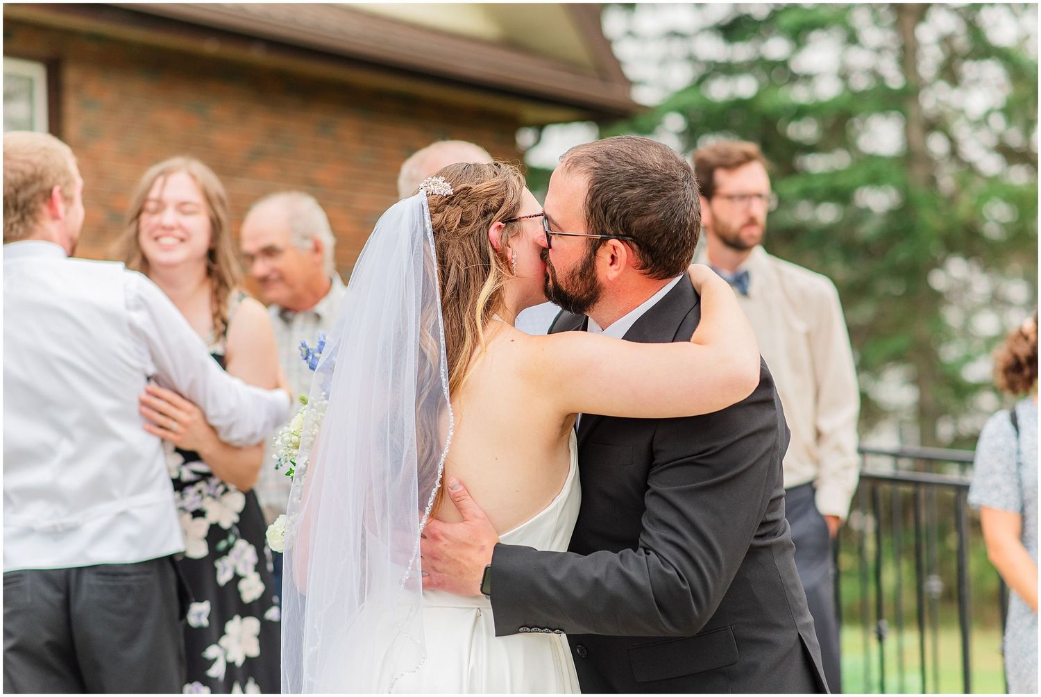 Newlywed couple sharing first kiss outdoors surrounded by cheering wedding guests after ceremony.