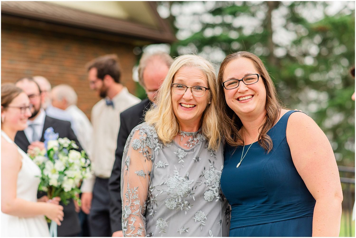Two women smile together outdoors at a wedding celebration, one in a silver beaded dress.