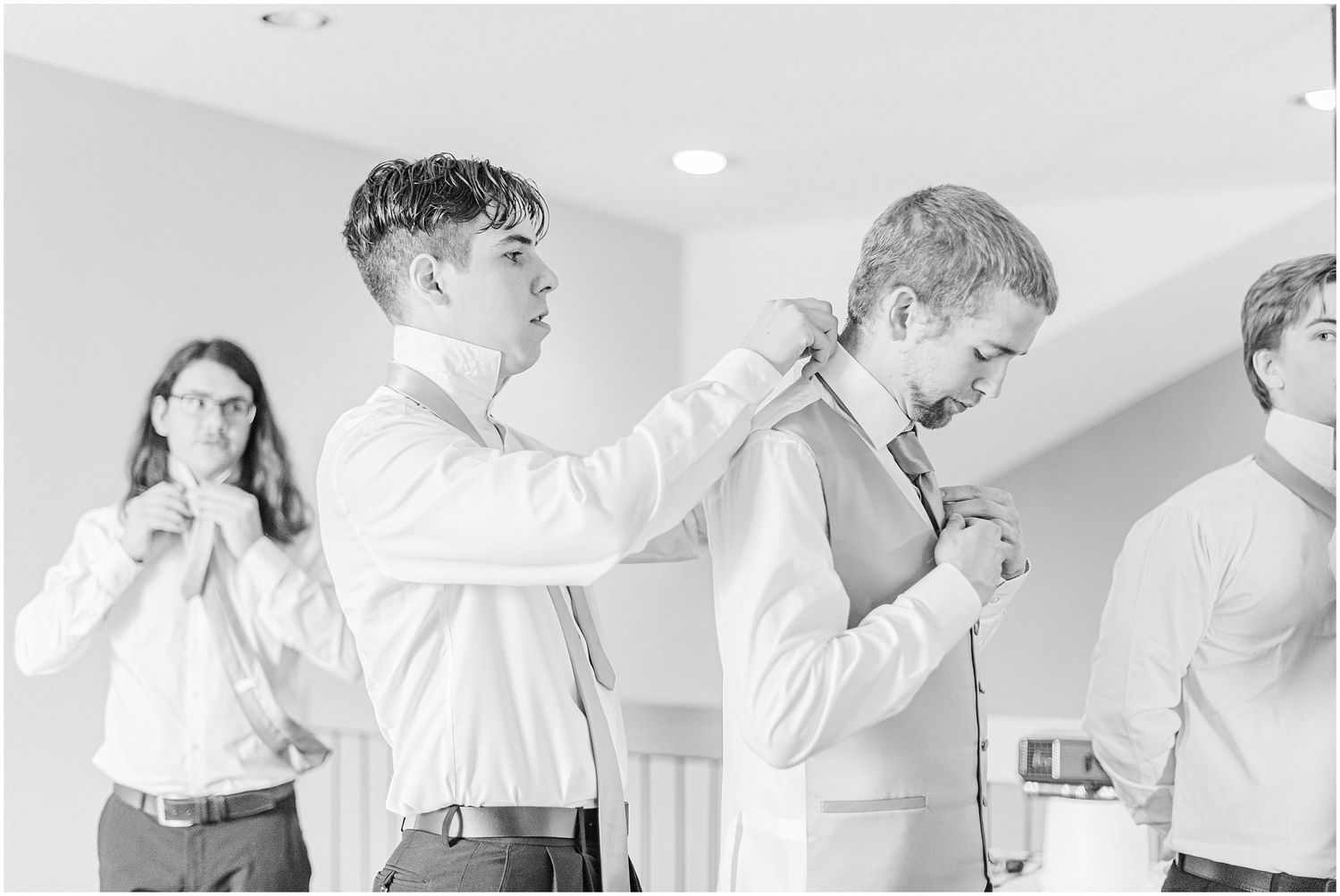 Groomsmen helping each other get dressed before a wedding ceremony in a bright room.