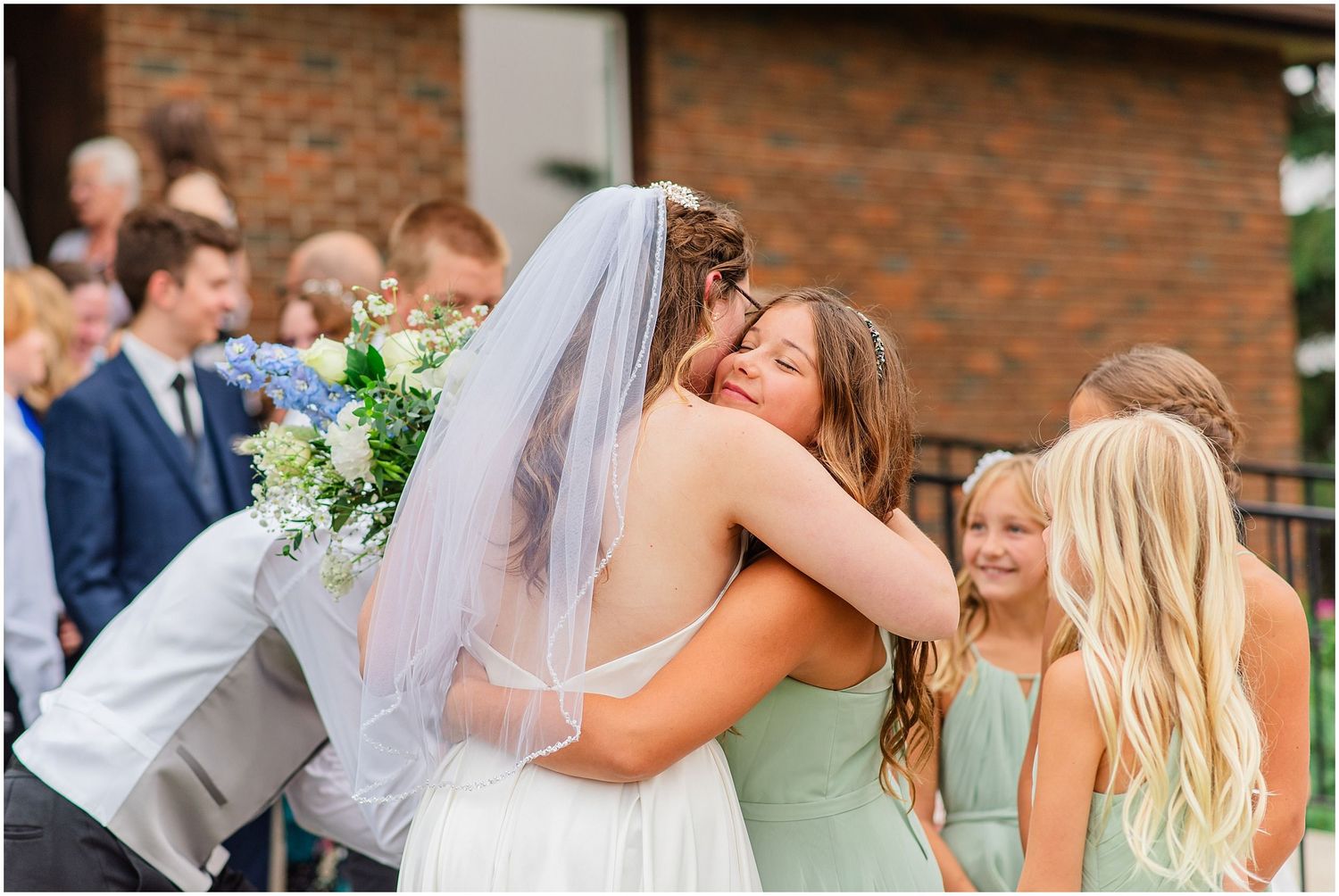 Bride in white gown and veil receiving warm hug from guest after outdoor wedding.
