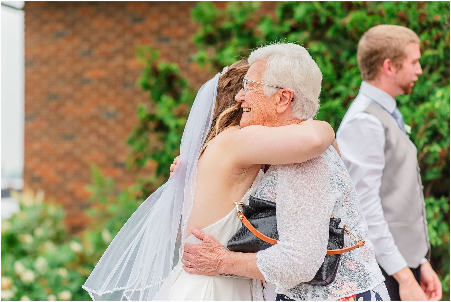 Bride in backless wedding gown embracing elderly guest in wheelchair after ceremony outdoors.