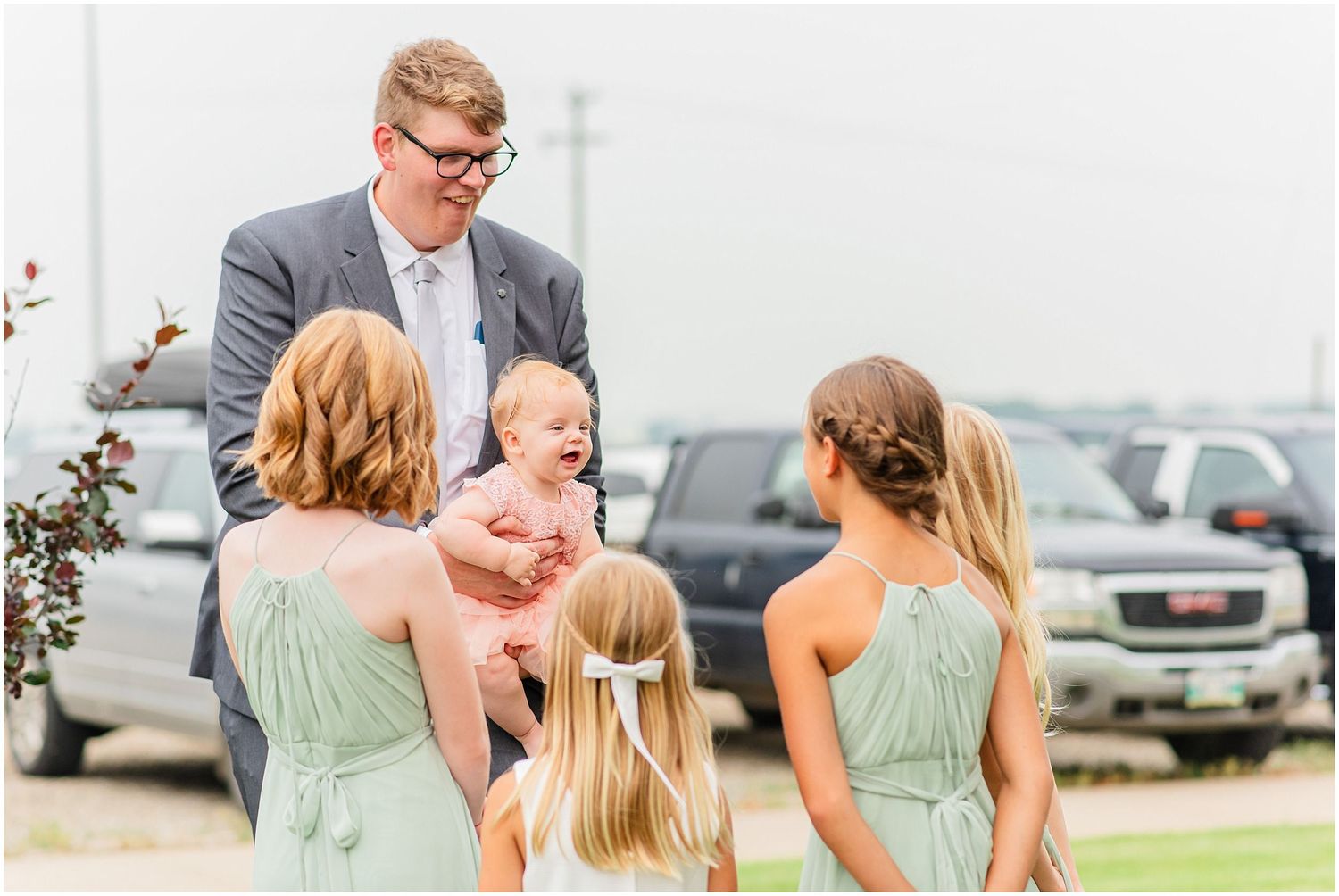 Groomsman in gray suit interacting with bridesmaids and a laughing baby outdoors at wedding.