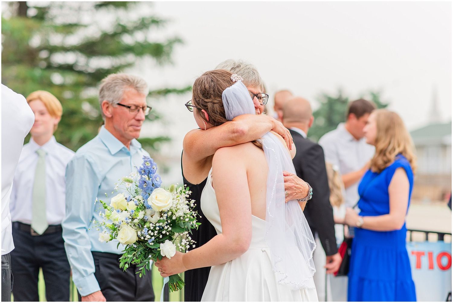 Bride embracing a guest at an outdoor wedding ceremony while holding a bouquet of white and blue flowers.