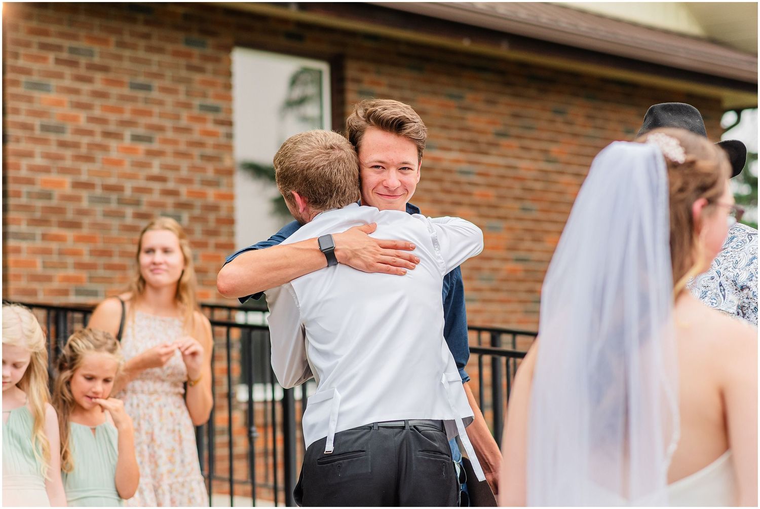 Groom smiling and hugging a guest outside a brick building during a wedding reception.