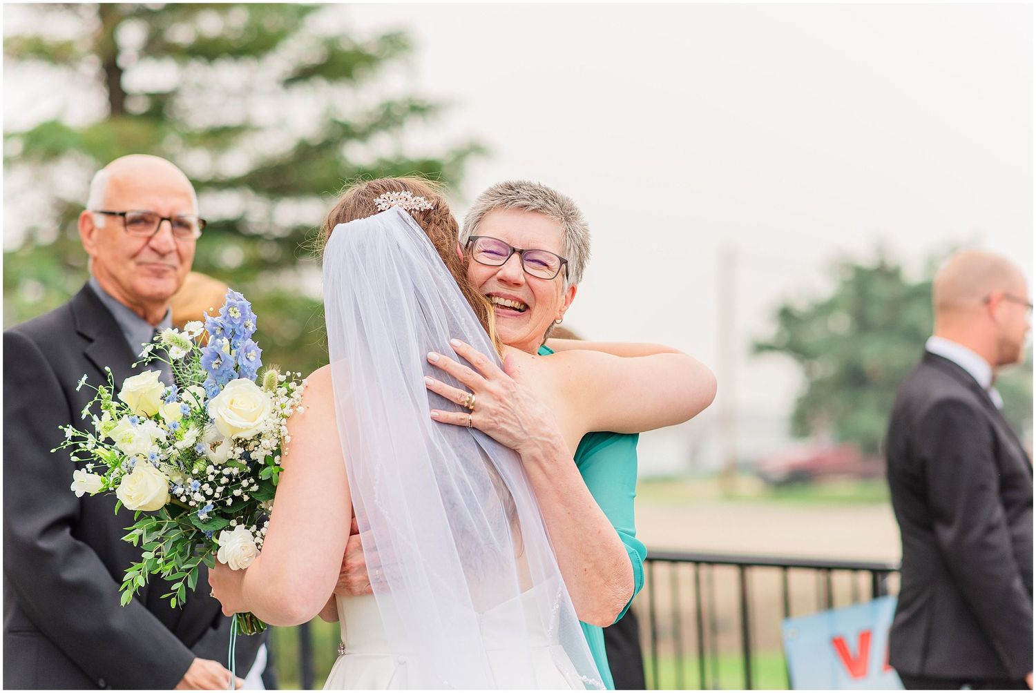 Bride in white veil receives warm hug from smiling older woman guest at outdoor wedding reception.