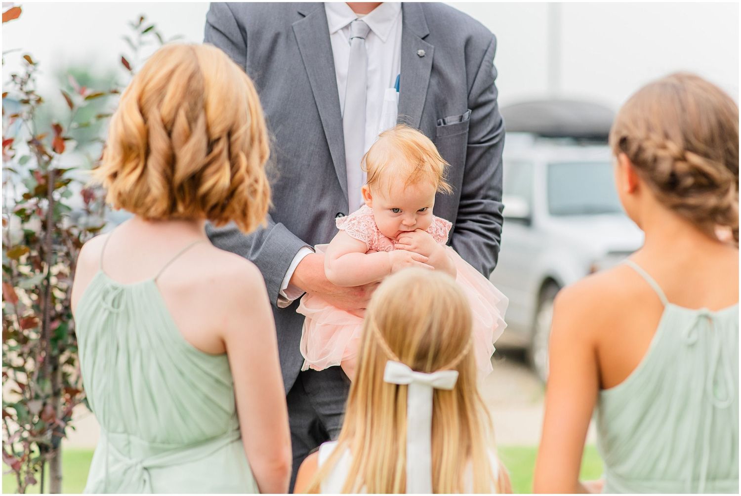Groomsman in gray suit holds baby in pink while flower girls in sage green dresses look on outdoors.