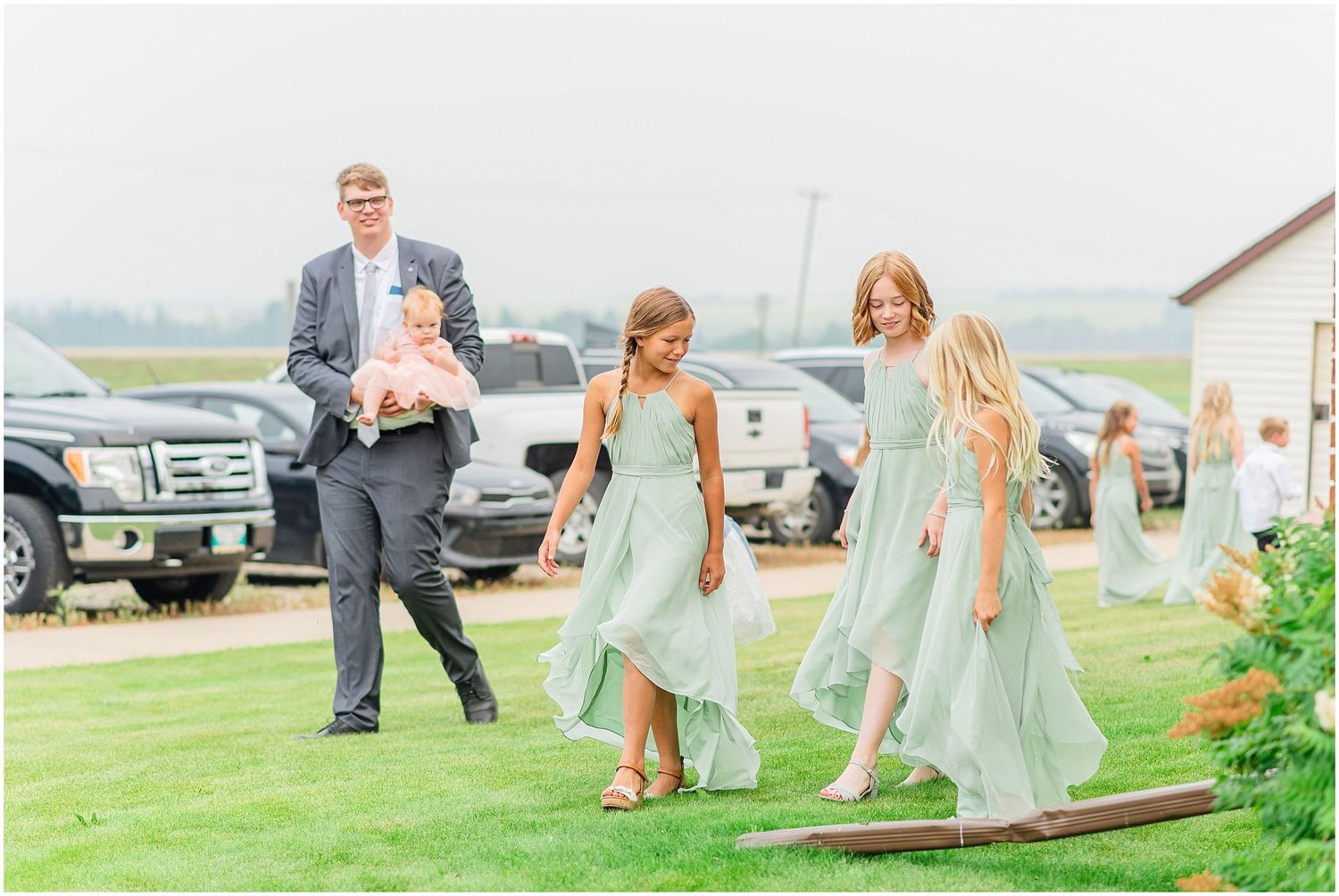 Groomsman carries baby while flower girls in sage green dresses walk across wooden plank at outdoor wedding.