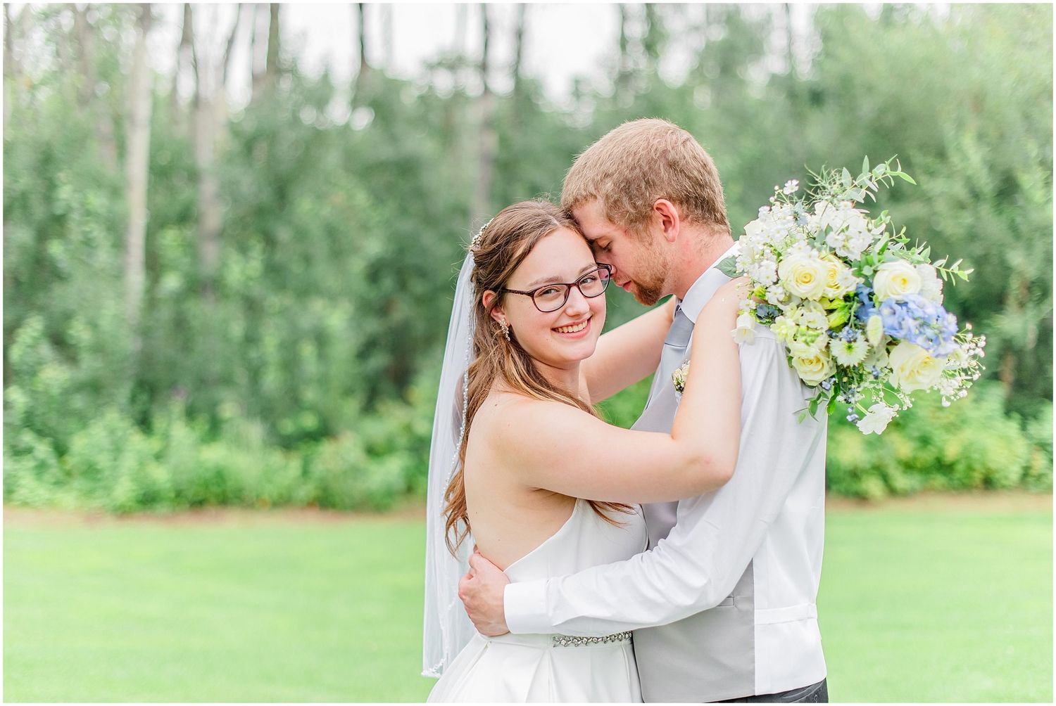 Smiling bride with glasses and groom touching foreheads outdoors holding blue and white wedding bouquet.
