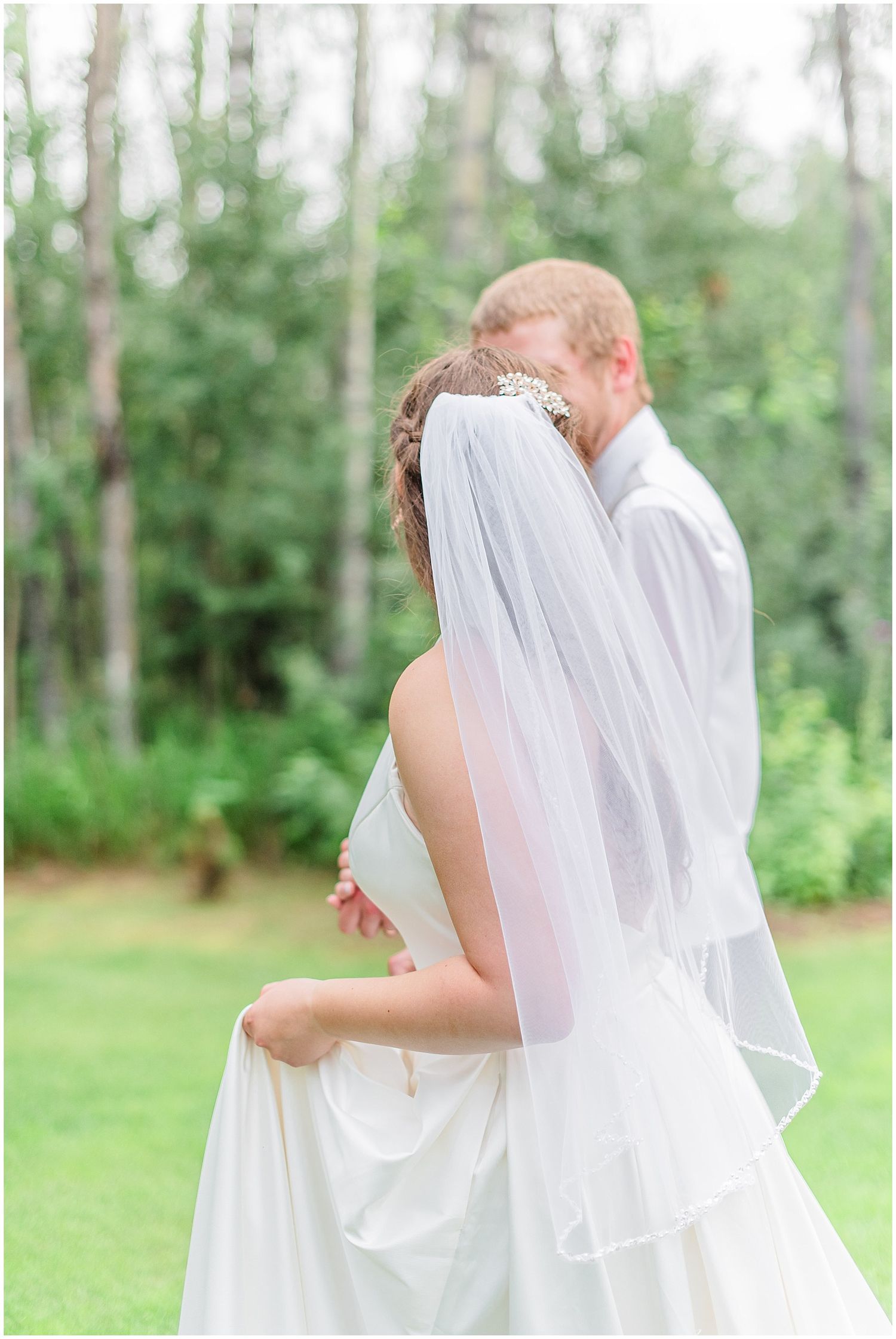 Bride and groom embracing from behind showing elegant white wedding dress and veil in lush green setting.