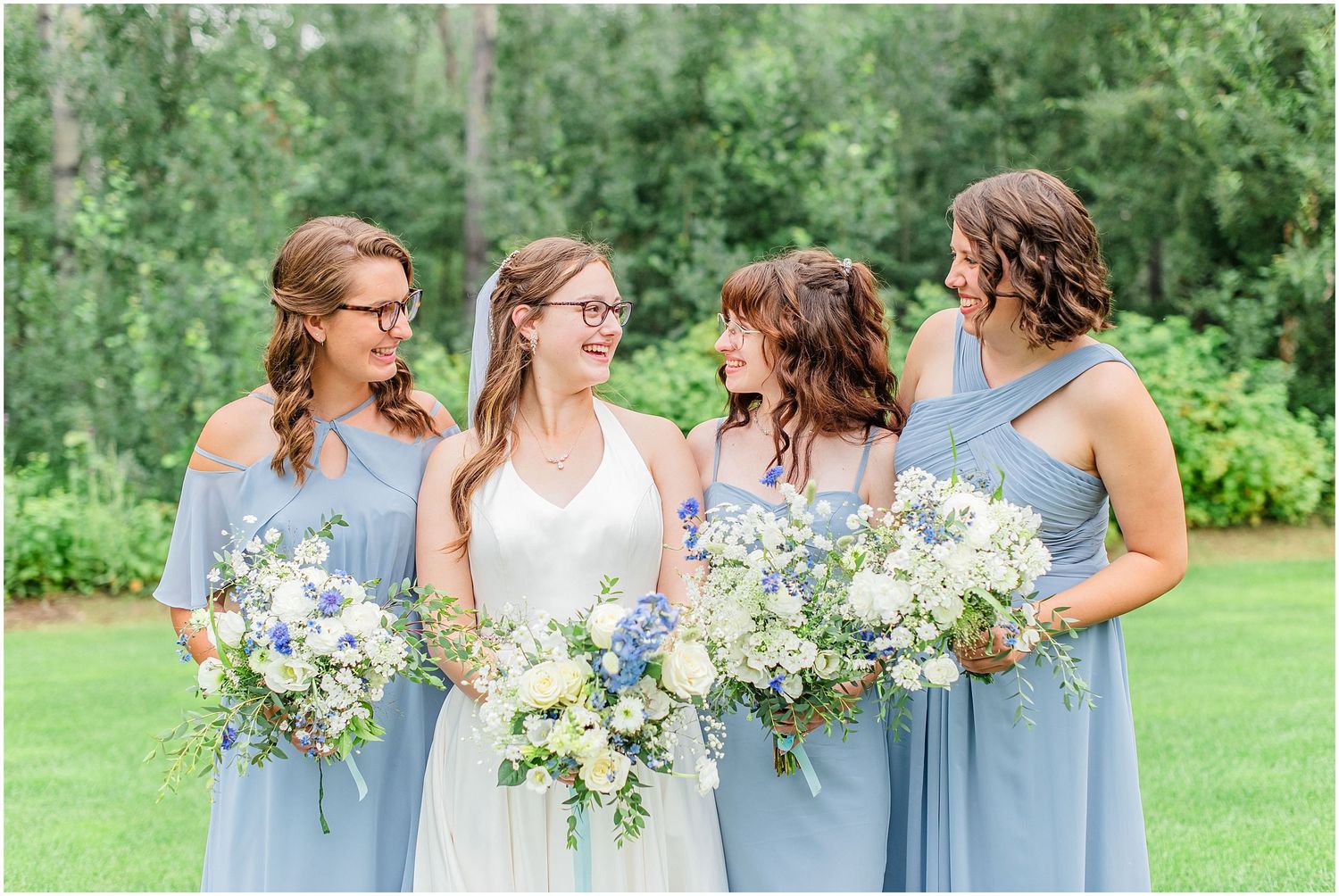 Smiling bride with bridesmaids in dusty blue dresses holding white and blue bouquets outdoors.