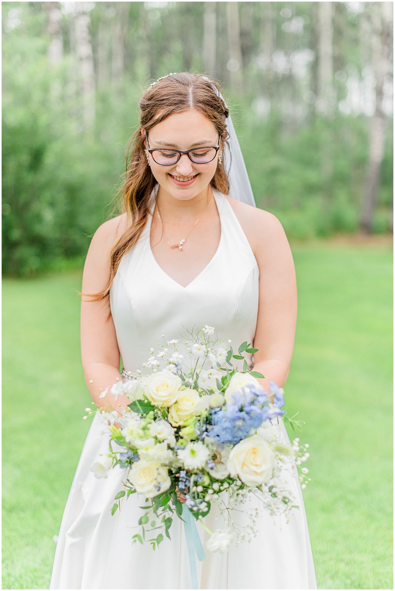 Bride in white halter gown smiles at her white and blue floral bouquet outdoors.