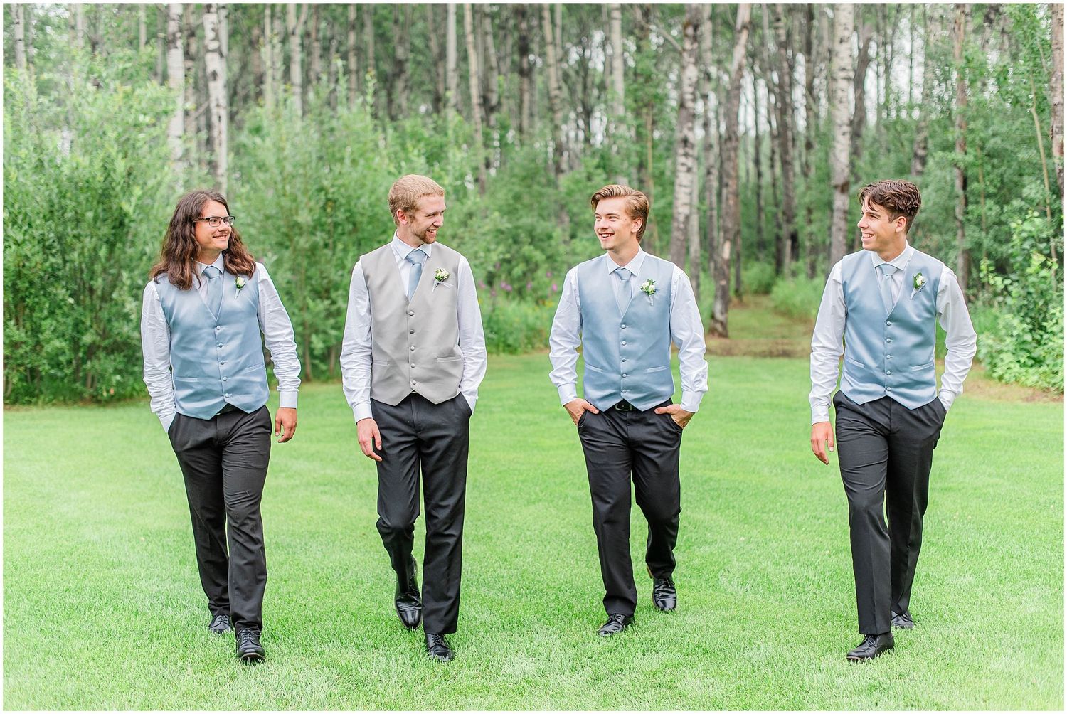 Four groomsmen in gray vests and blue shirts walk together across green lawn at outdoor wedding.