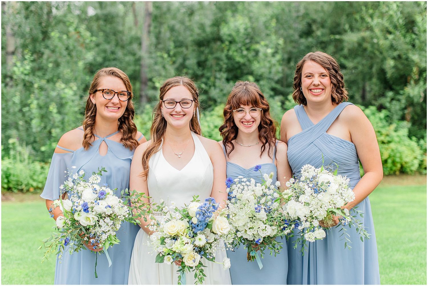 Bride with three bridesmaids in dusty blue dresses holding white and blue floral bouquets outdoors.