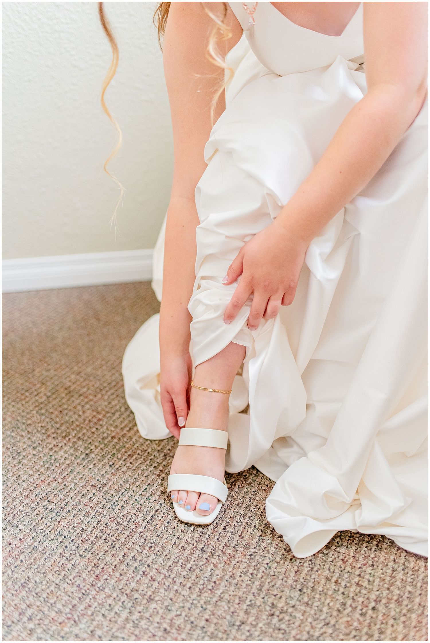 Bride in white gown adjusting her white block heel sandal while seated on carpet before wedding ceremony.