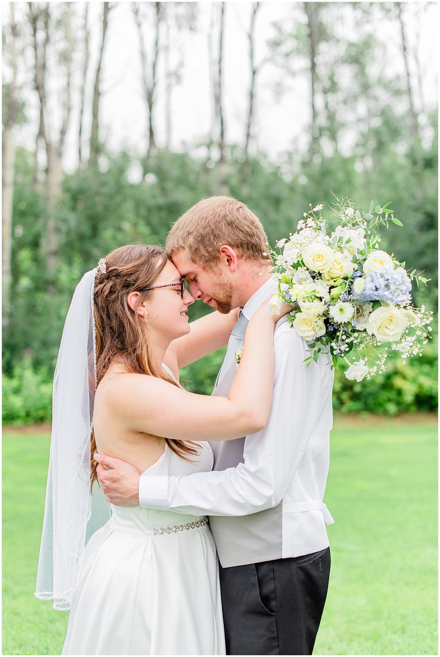 Bride and groom touch foreheads tenderly while bride holds white and blue floral bouquet outdoors.