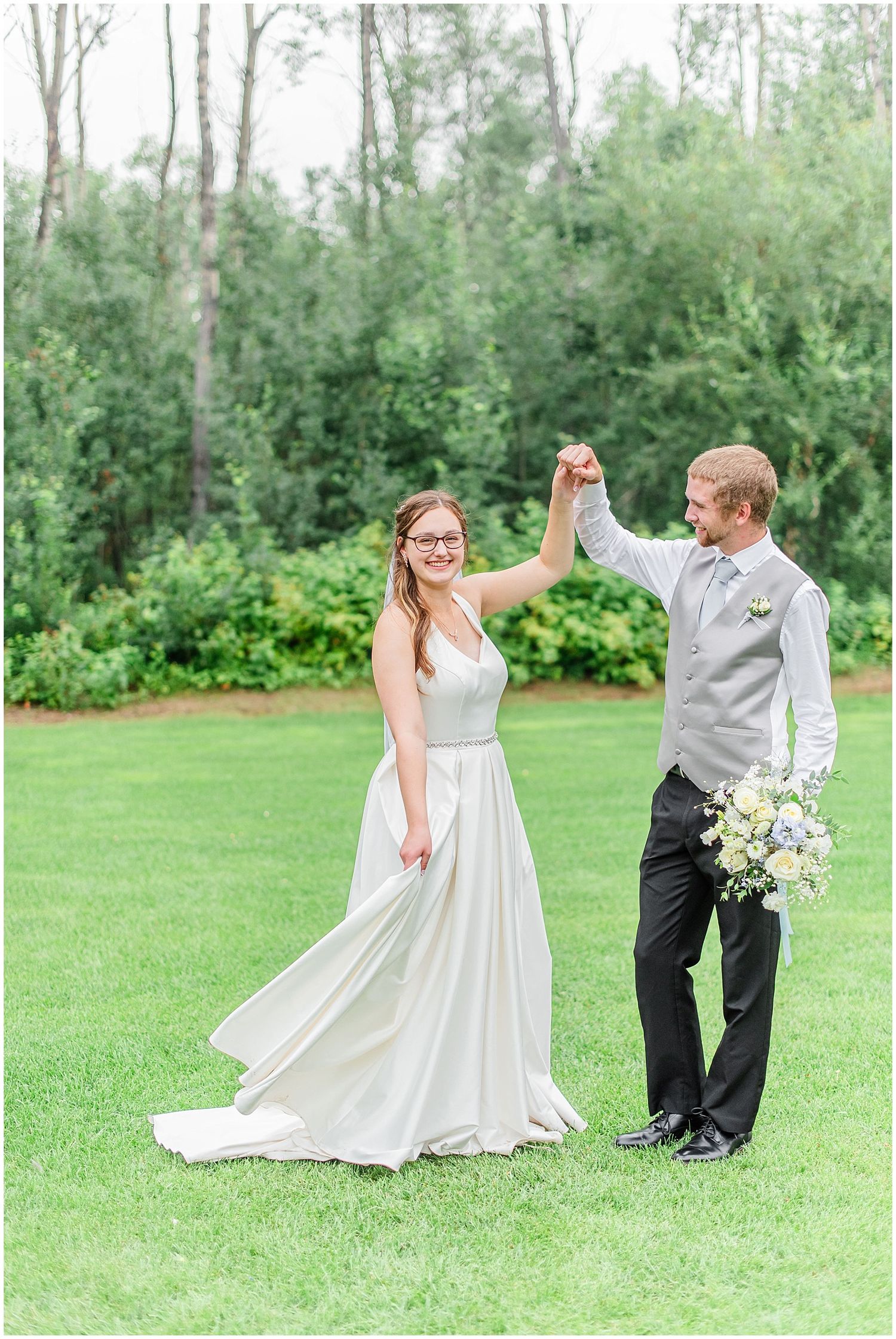 Groom twirls his bride in a flowing white gown on a lush green lawn surrounded by trees.