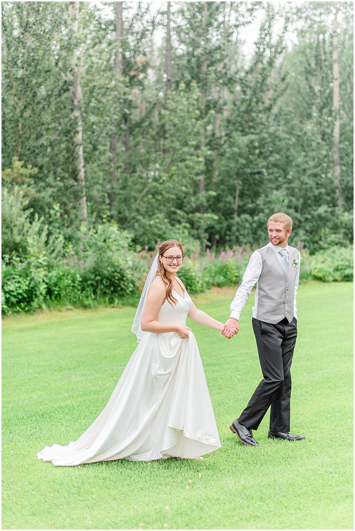 Smiling bride in white gown and veil holds grooms hand walking across lush green lawn.