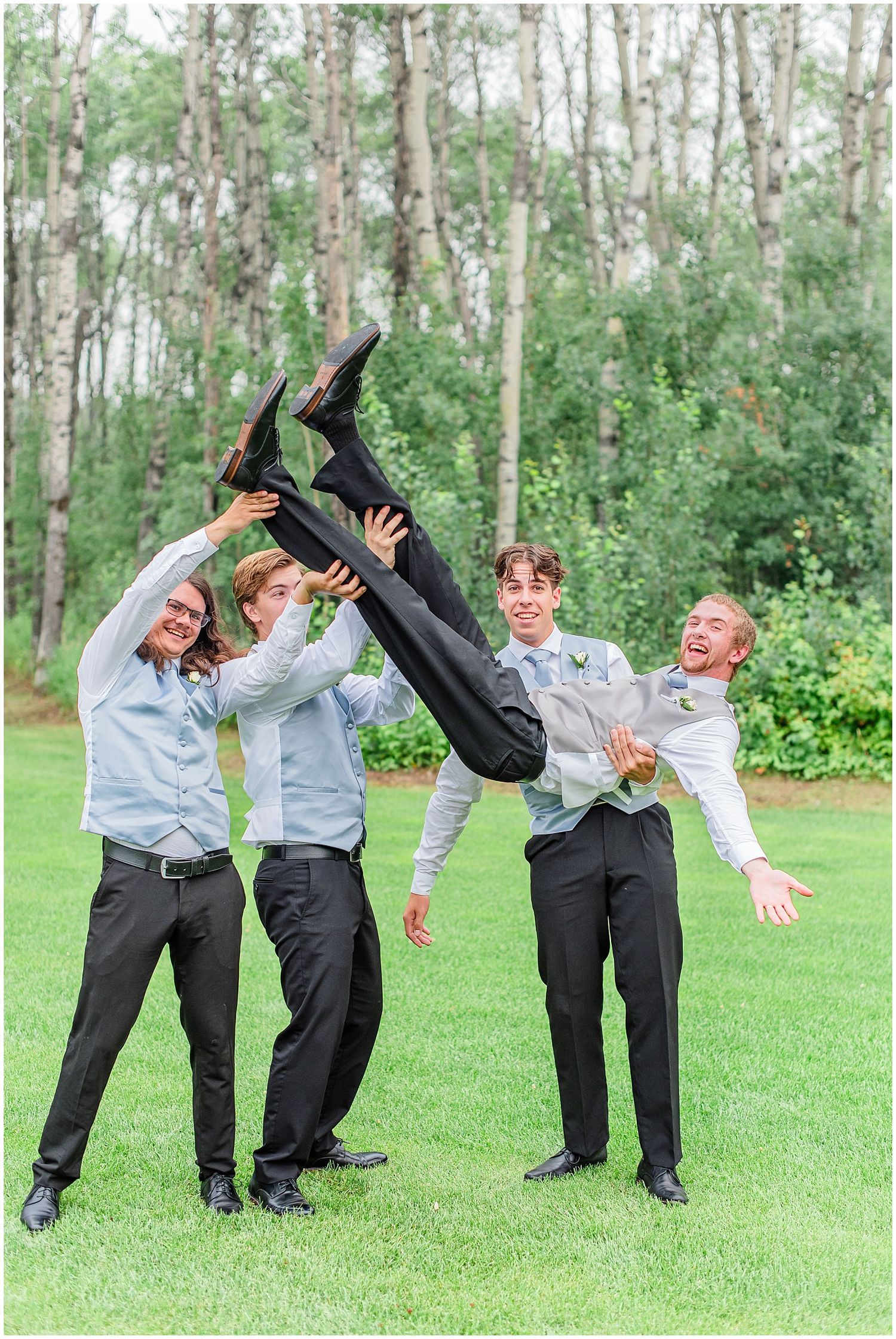 Groomsmen playfully lift the groom in the air on a green lawn with trees behind them.