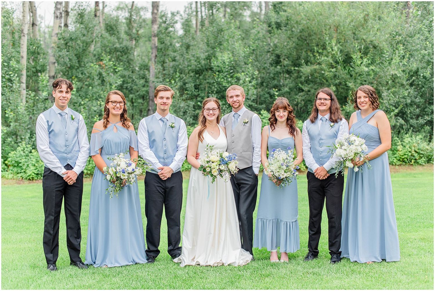 Wedding party in dusty blue dresses and gray suits poses together outdoors near lush green trees.