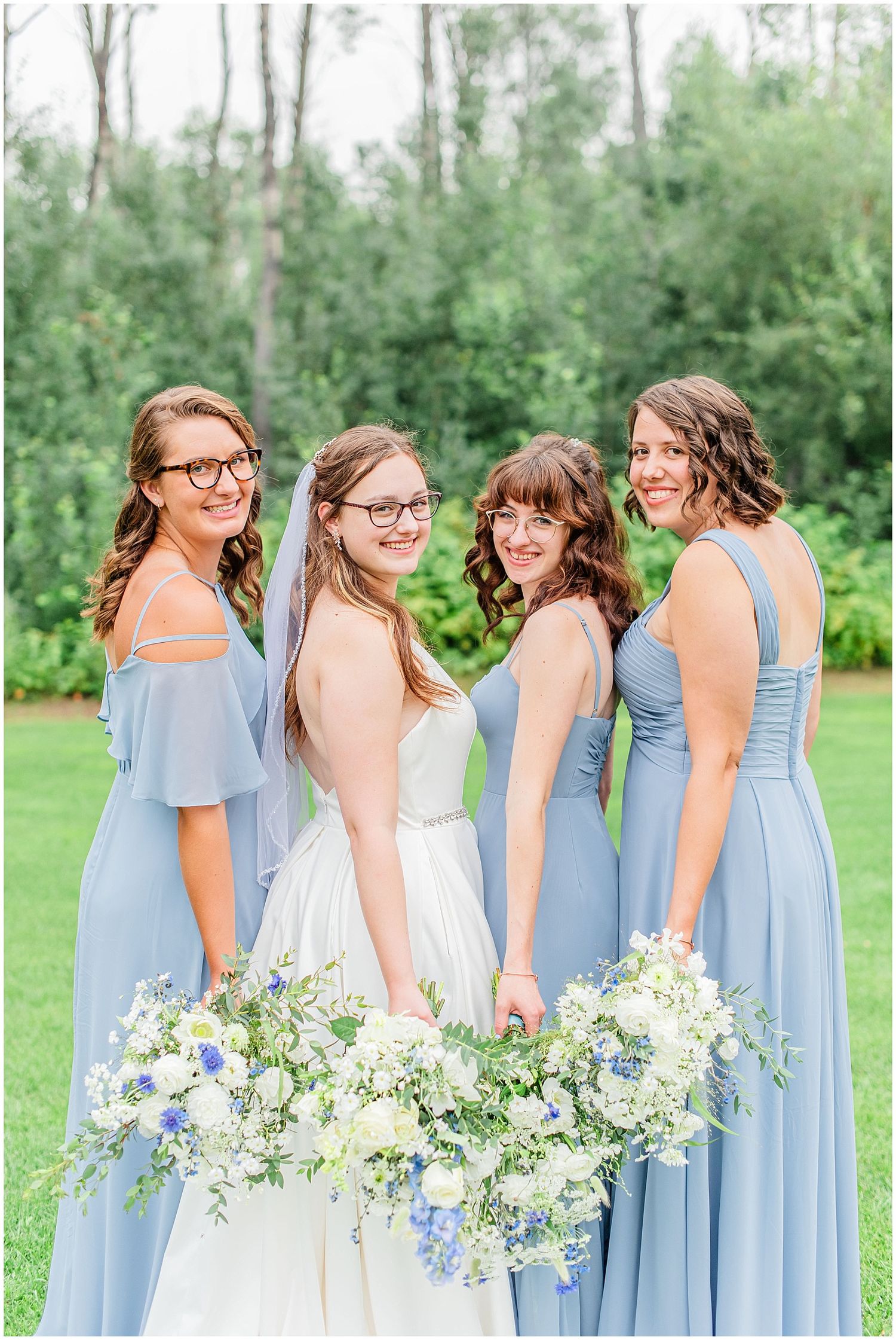 Bride in white gown poses with three bridesmaids in dusty blue dresses holding white and blue floral bouquets outdoors.