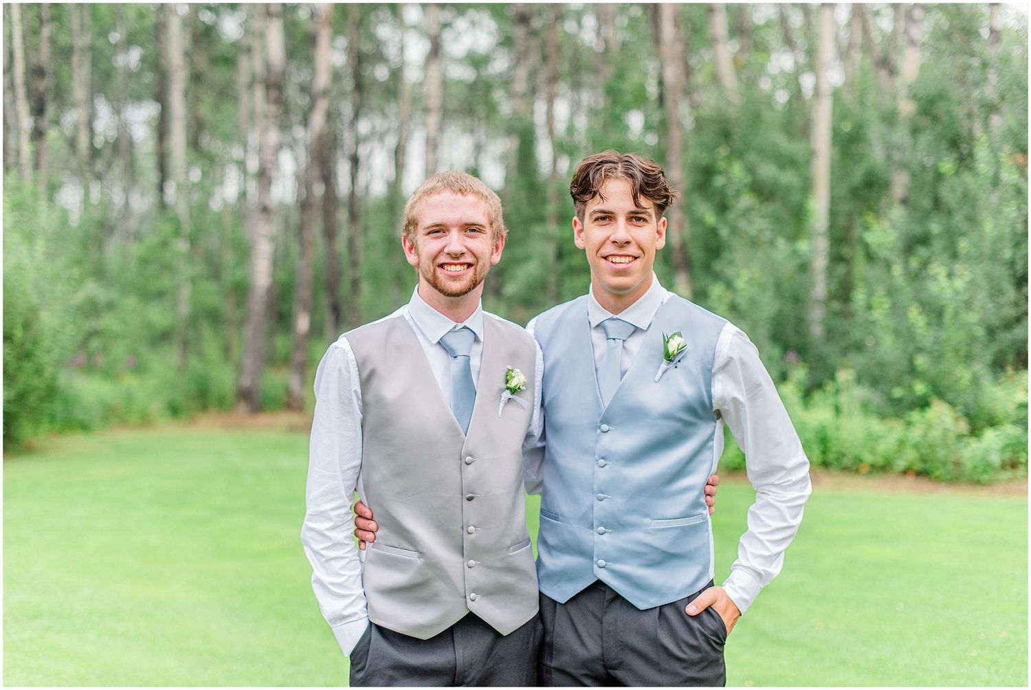 Two groomsmen smile in gray vests and blue ties with boutonnieres on a green lawn.