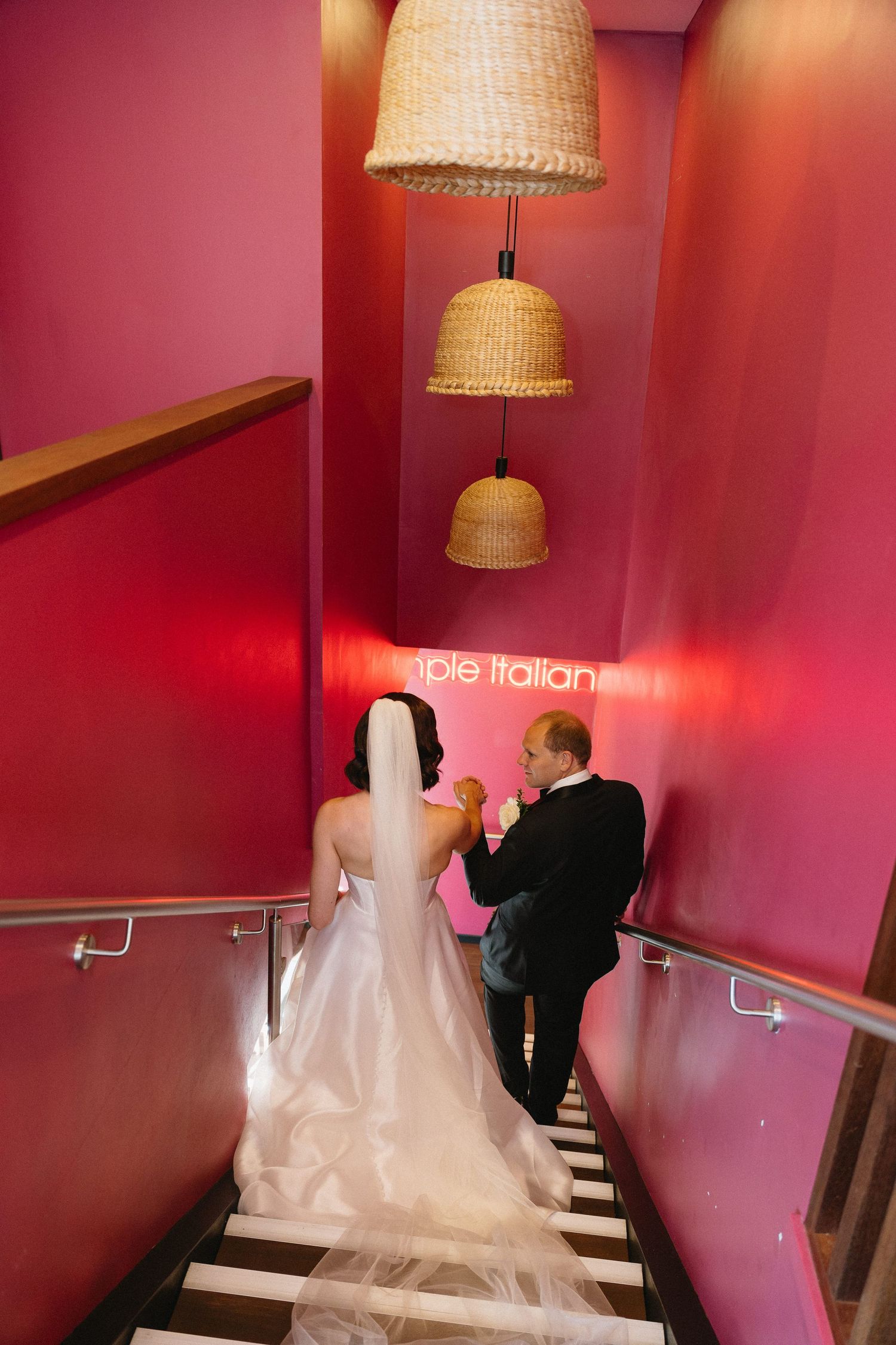 Bride and groom descend vibrant pink staircase with wicker pendant lights and neon sign at elegant wedding venue.