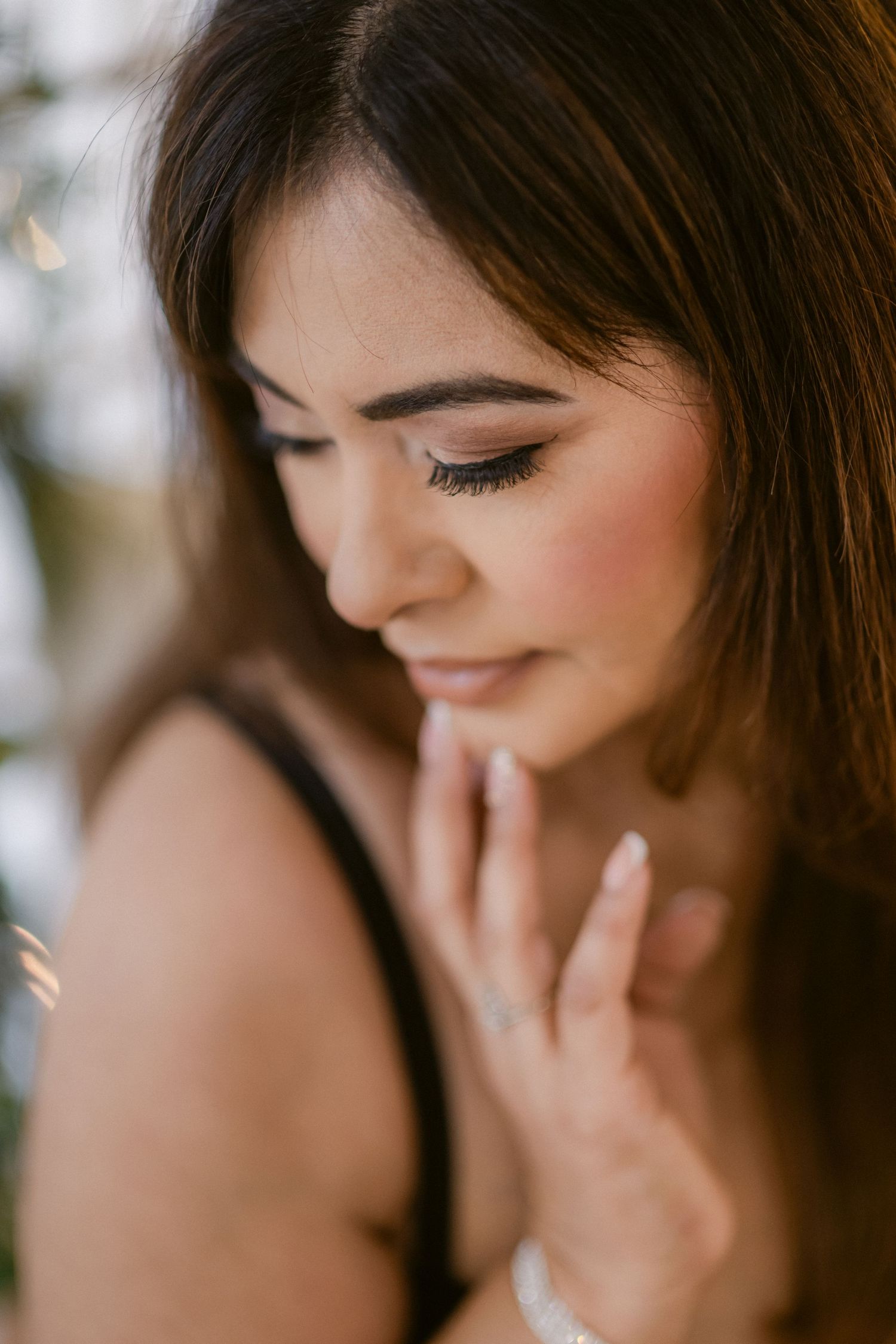 Close-up portrait of a brunette woman with elegant makeup, looking down gracefully with hand near her face.