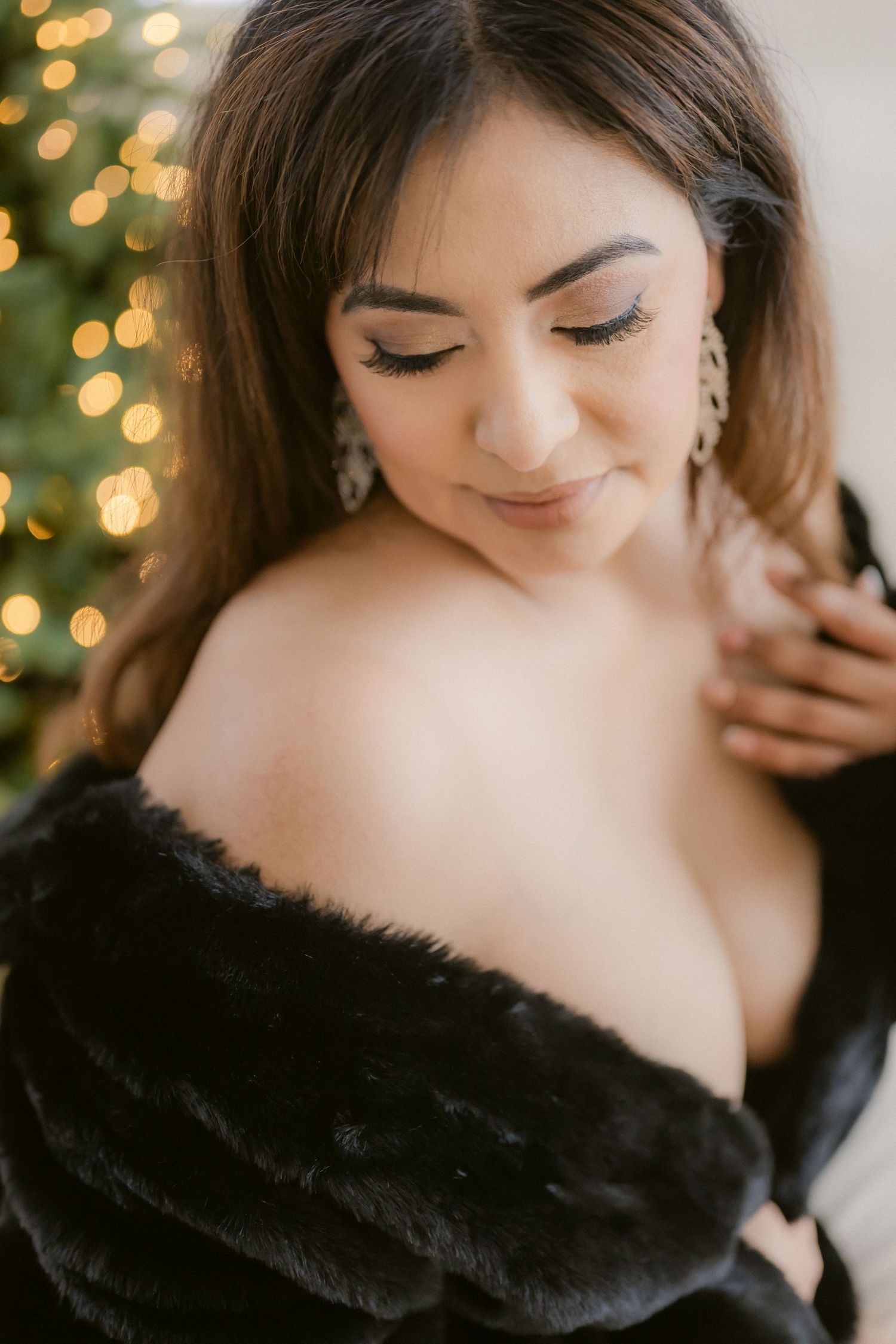 Woman in black off-shoulder fur coat with diamond earrings posing near Christmas tree bokeh lights.