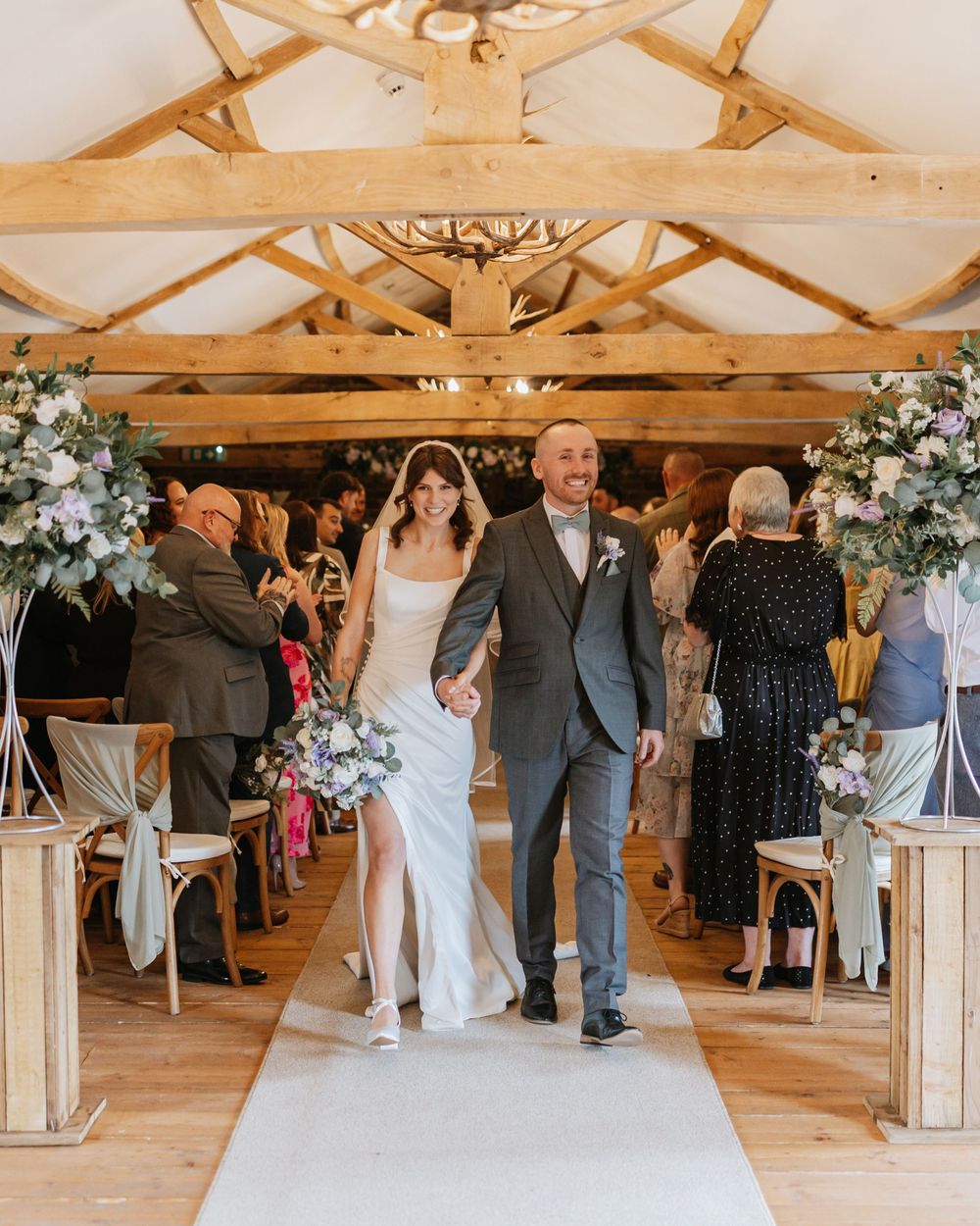Bride and groom walk down the aisle in a rustic barn wedding venue with wooden beams and floral arrangements.
