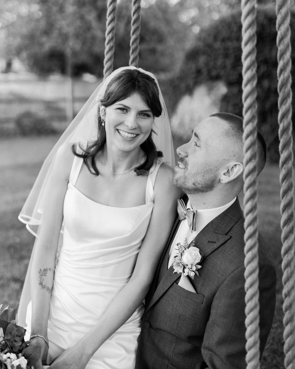 Black and white photo of a smiling bride in a veil and groom in a suit with boutonniere on a rope swing.