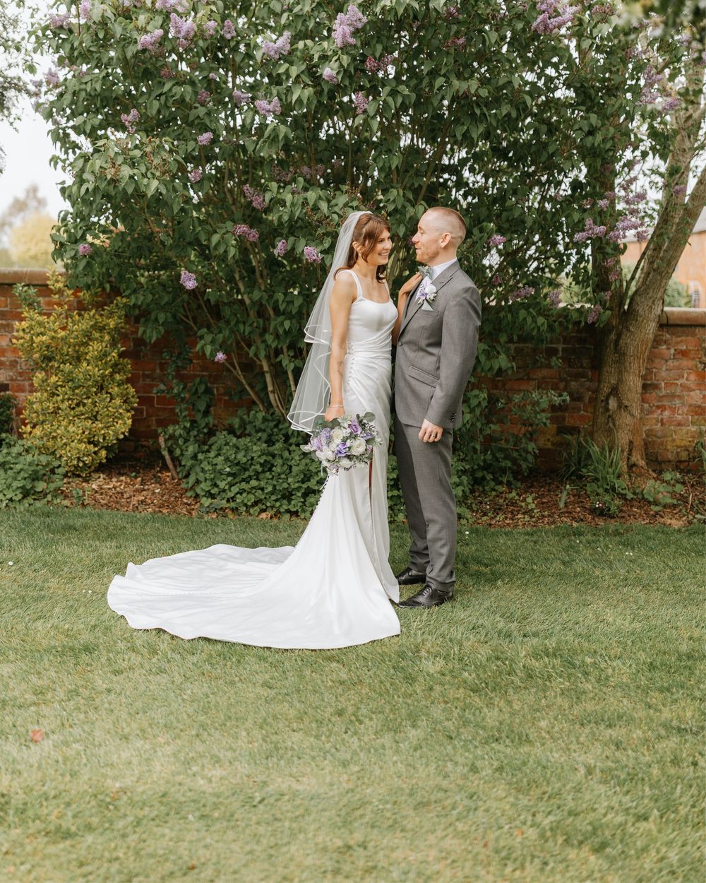 Bride and groom stand on green lawn before blooming lilac tree, she in white gown with veil, he in gray suit.