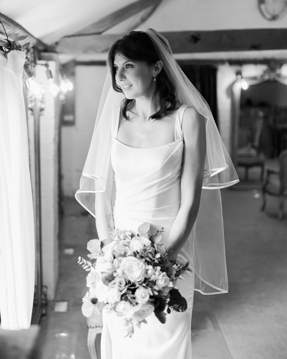 Black and white photo of a bride in a sleek white gown and veil holding a floral bouquet indoors.
