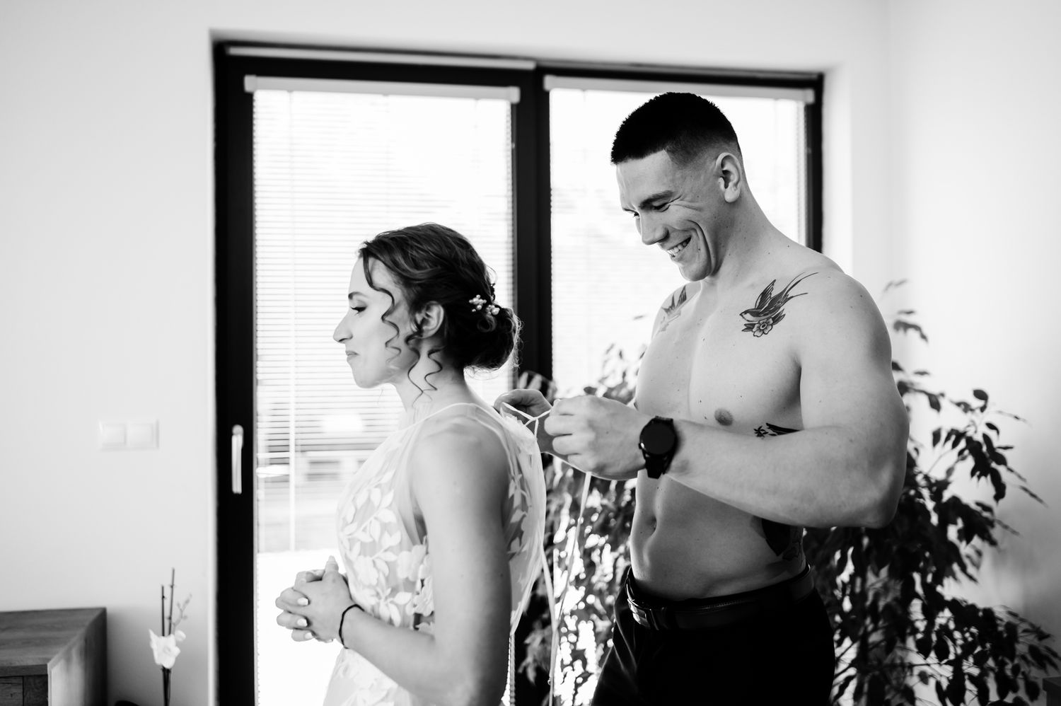 Bride in white lace gown stands beside groom in white shirt during wedding preparation indoors.