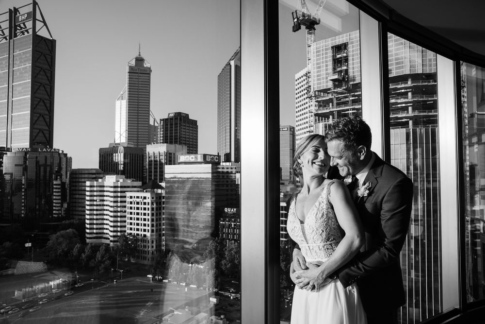 Black and white wedding portrait of couple embracing by floor-to-ceiling windows with city skyline backdrop.