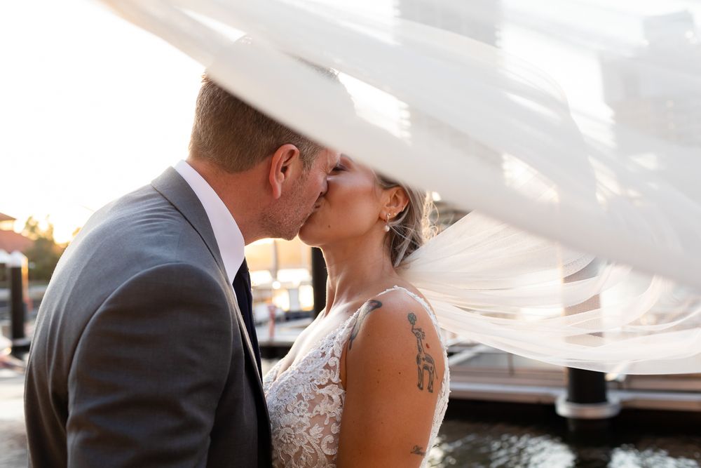 A bride and groom share a romantic kiss as her veil billows in the wind near a waterfront venue.