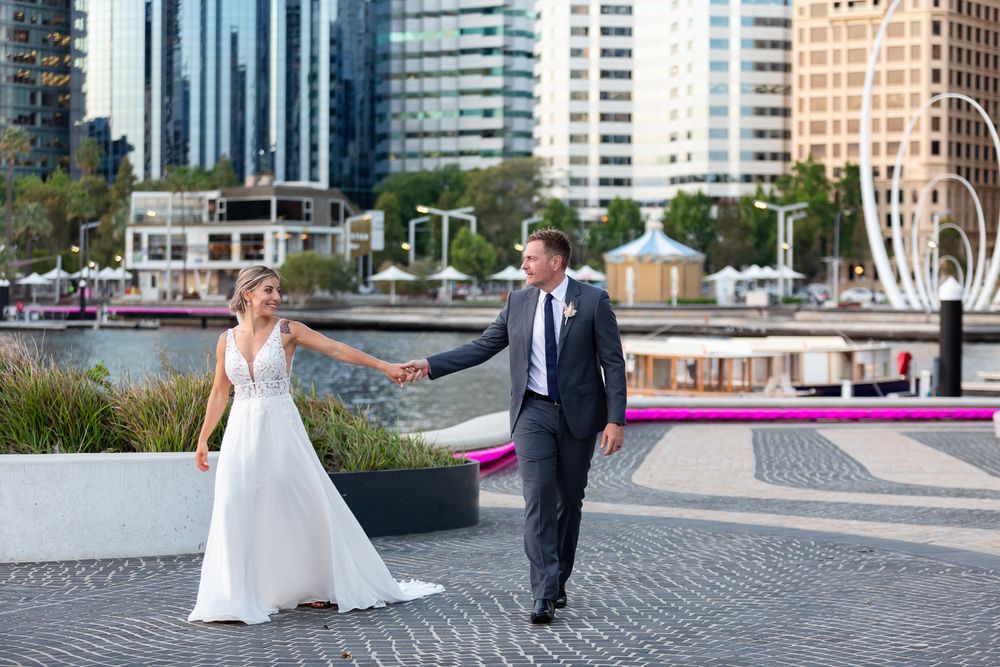 Bride and groom hold hands walking along a waterfront plaza with city skyscrapers in the background.