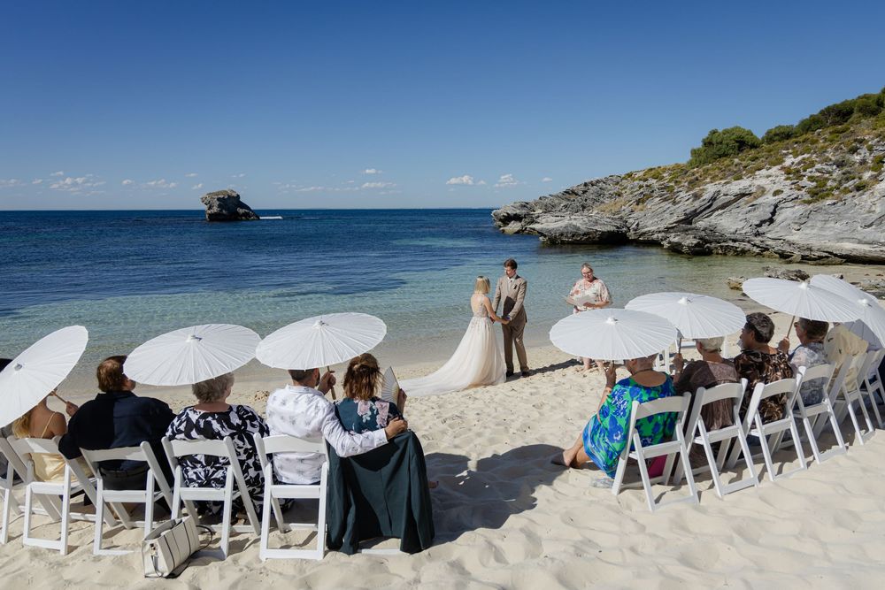 Beach wedding ceremony with guests seated in white chairs under parasols, couple exchanging vows by the ocean on a sunny day.