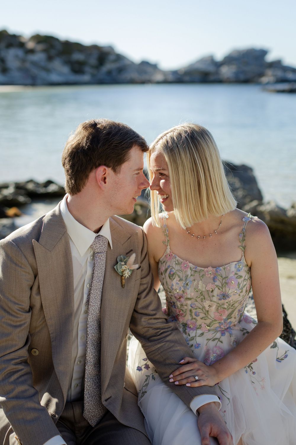 Couple in wedding attire sharing a romantic moment by rocky coastal waters, bride in floral dress, groom in tan suit.