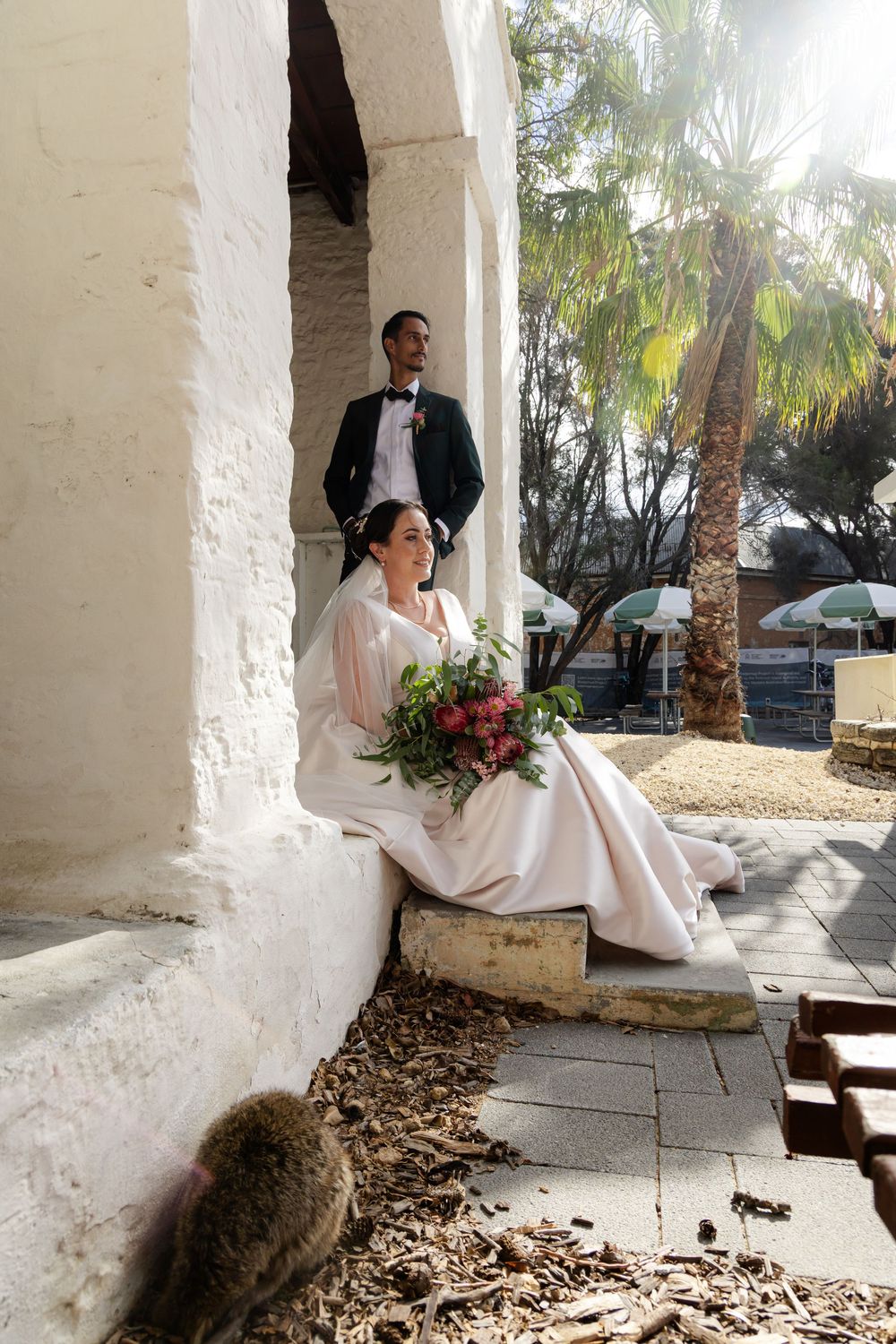 Bride sits on steps holding red bouquet while groom stands behind her near a sunlit palm tree.