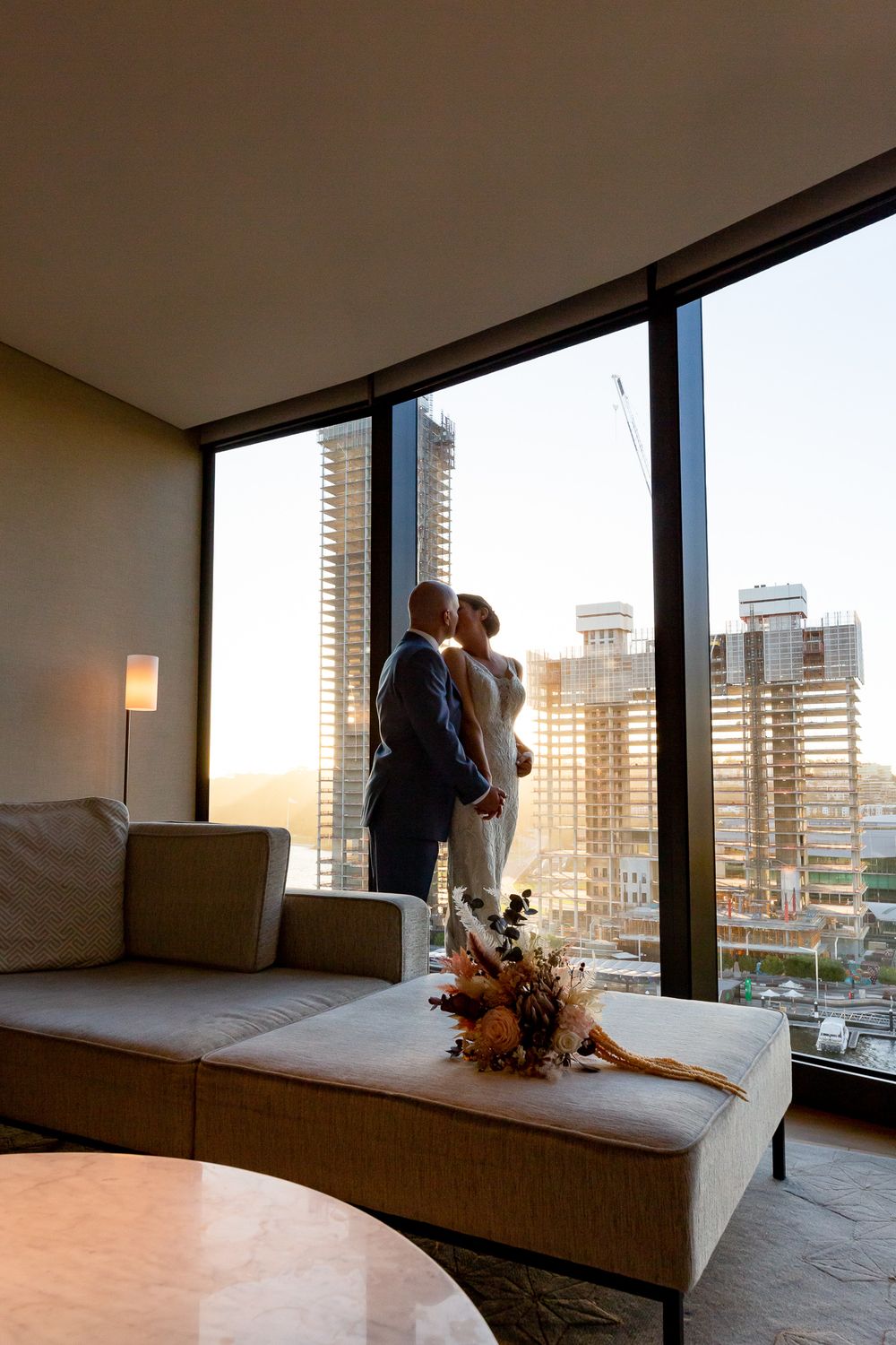Newlyweds kiss by floor-to-ceiling windows with city skyline at golden sunset.
