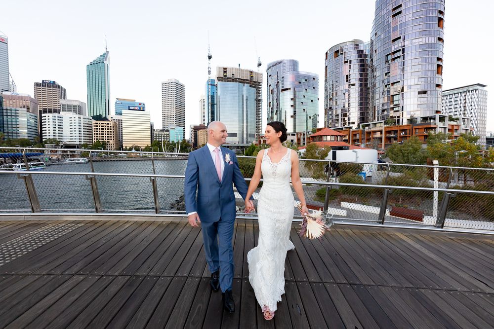 Bride and groom walking hand in hand on a waterfront boardwalk with a city skyline backdrop.
