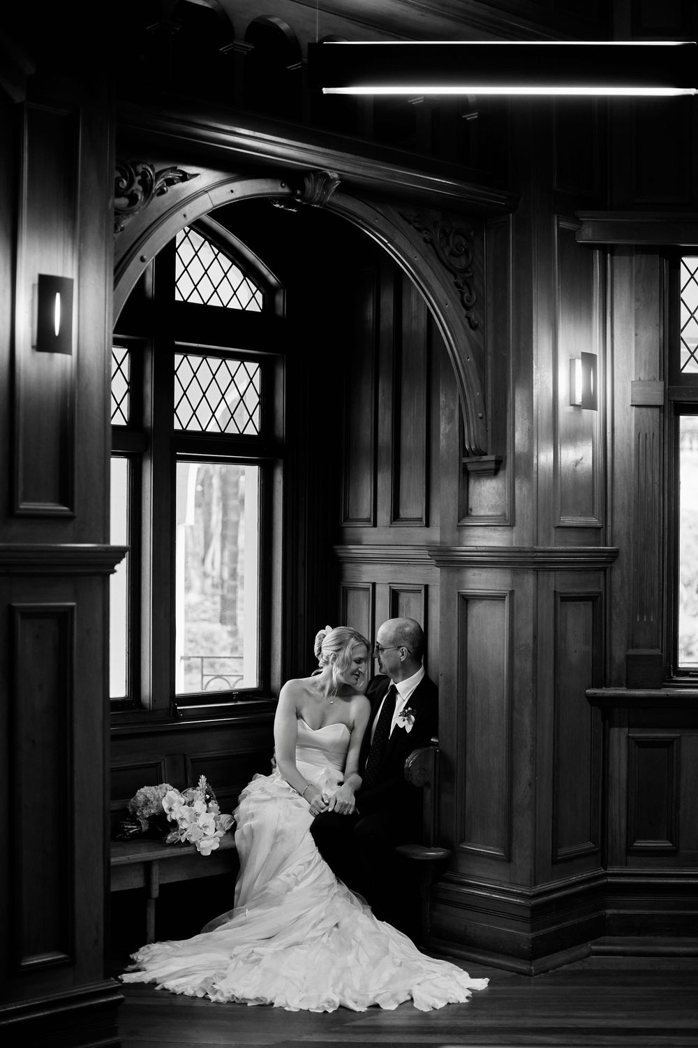 Black and white wedding portrait of bride and groom sitting together in elegant wood-paneled room with arched windows.