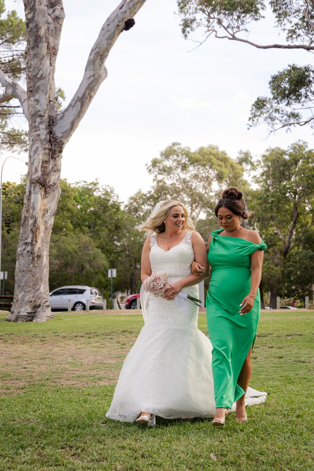 Bride in white gown walks with bridesmaid in green dress across garden lawn at outdoor wedding ceremony.
