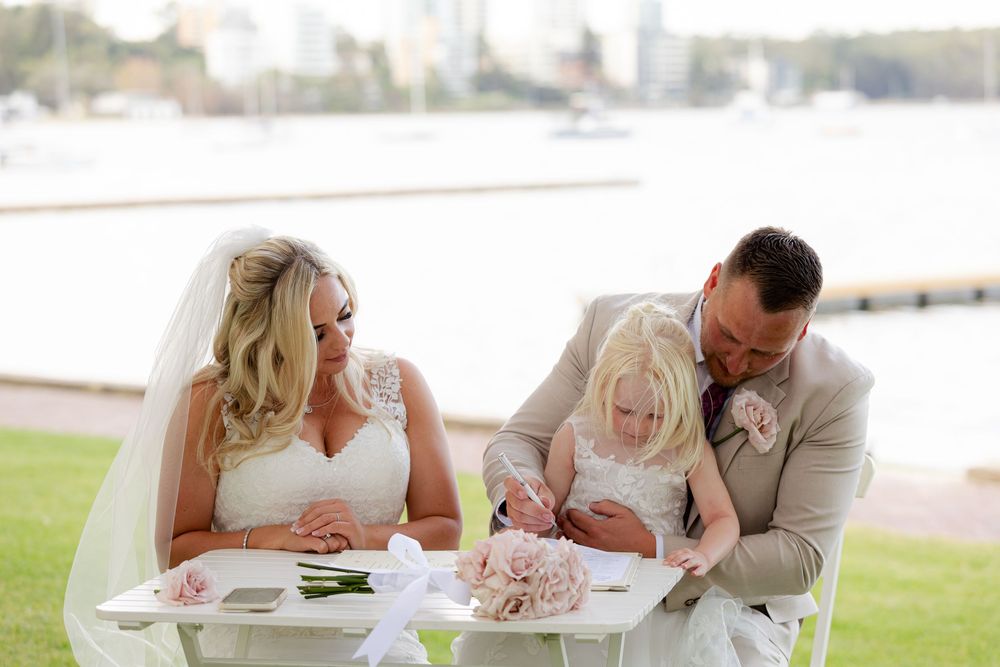 Bride and groom signing wedding documents outdoors with young flower girl at white table near waterfront.