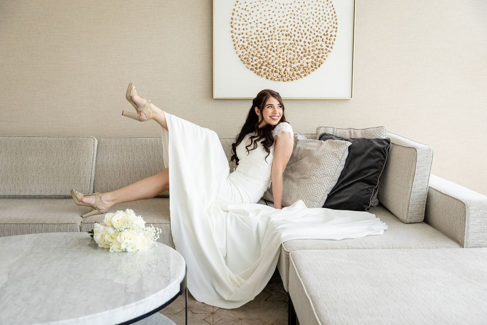 Bride in white gown posing elegantly on a gray sofa with heels and a bouquet nearby.