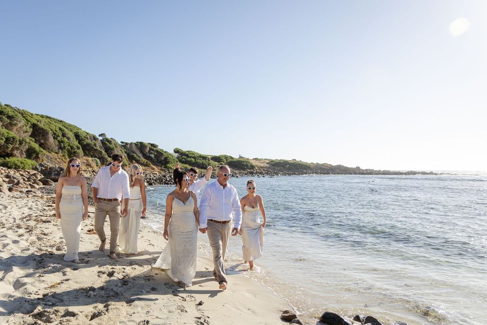 Wedding party walking along a sunny beach in white and beige attire with blue ocean and green hills backdrop.