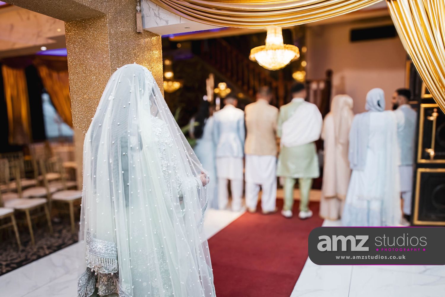 A veiled South Asian bride stands before a red-carpeted aisle during a traditional wedding ceremony indoors.
