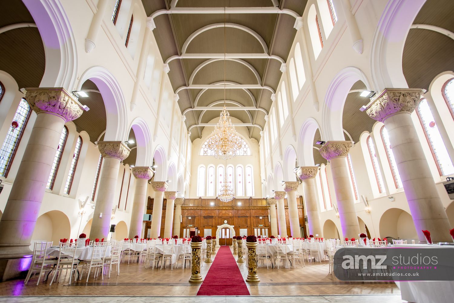 Elegant church wedding venue with red carpet aisle, white chairs, chandelier, and purple uplighting in a grand vaulted hall.